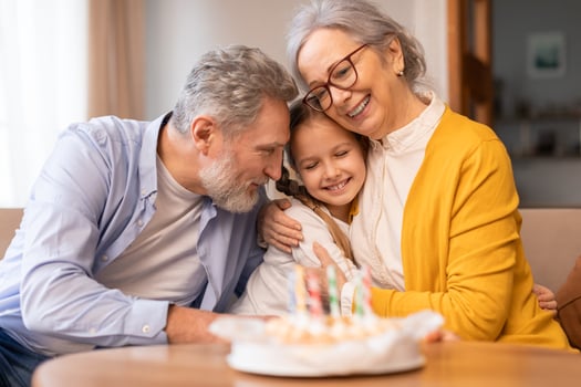 Senior man and woman are sitting next to a little girl in front of a birthday cake. They seem to be celebrating a special occasion, possibly a birthday, with the cake as the centerpiece photo