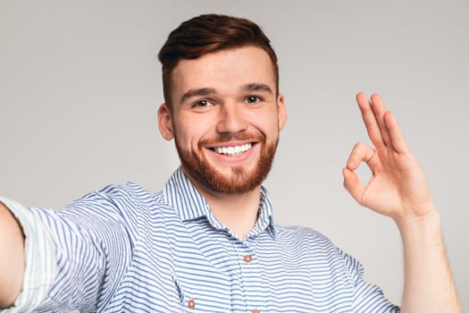 Handsome redhaired young guy showing OK sign and making selfie against grey studio background, closeup photo