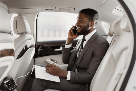Black businessman is sitting in the backseat of a car, taking notes on a notepad while talking on a phone. He is wearing a suit and tie, and he has a serious expression on his face. photo