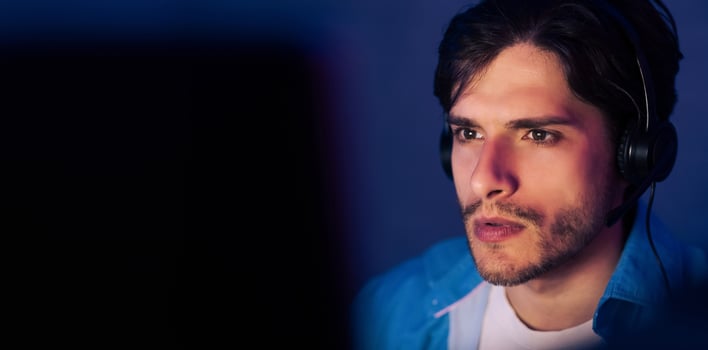 A young man with dark hair is wearing headphones and looking intently at a computer screen. He is wearing a blue jacket and a white t-shirt. The image is taken from a close-up perspective, copy space photo
