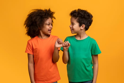 This image features two young African American children, a girl wearing an orange shirt and a boy wearing a green shirt, smiling and holding pinky fingers in front of a yellow background. photo