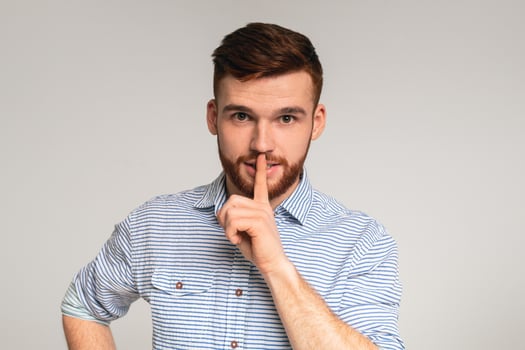Do not speak. Young man showing silence sign and putting finger to lips on studio background, closeup shot photo