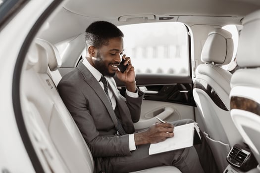 African American businessman in a suit sits in the backseat of a luxury car and makes notes on a notepad while speaking on his phone. He looks focused and professional. photo