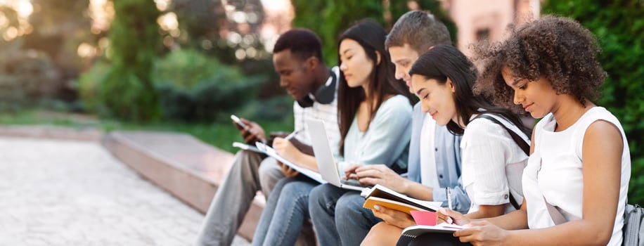 Educational Process. Group Of College Students Studying Outdoors, Sitting On Bench In Campus With Workbooks And Laptop, Side View photo