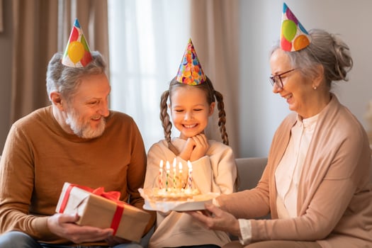 In a warm, cozy room, a young girl is delighted with a lit birthday cake presented by her smiling grandmother, while a happy grandfather looks on and holds a wrapped present photo