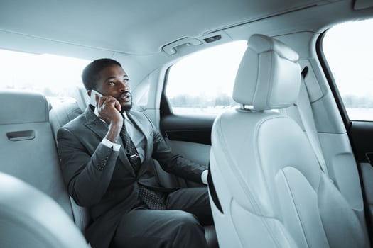 African American businessman, dressed in a black suit and tie, is sitting in the back seat of a car and talking on his phone. He is looking out the window as he speaks photo