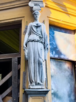 A close-up view of a classical female statue mounted on the corner of a historic building, illuminated by warm sunlight. The weathered stone figure contrasts with the vibrant yellow facade photo