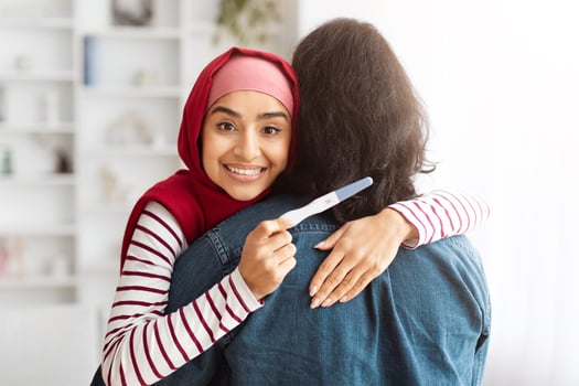 A young muslim woman wearing a red hijab expresses happiness as she warmly embraces her husband, holding positive pregnancy test photo