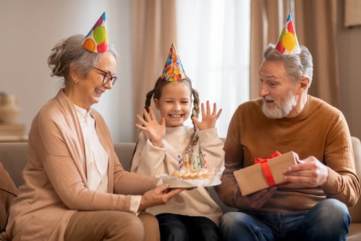 A young girl wearing a party hat is sitting between her smiling grandparents, who are also adorned with colorful party hats, grandfather is excitedly giving her birthday present wrapped in red paper photo