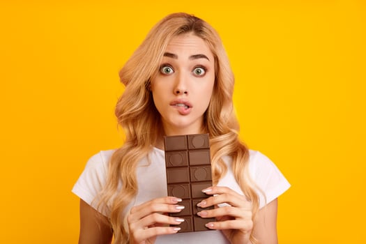 A young woman with long, wavy blonde hair is holding a chocolate bar in front of her face. She is wearing a white t-shirt and is looking at the camera with a surprised expression photo