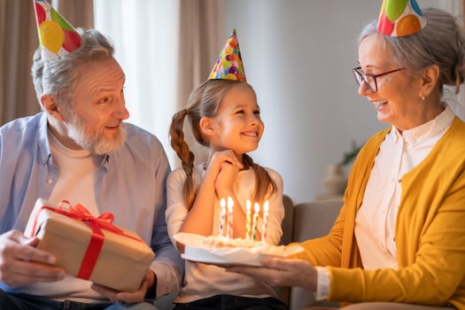 A young girl clad in a birthday hat smiles brightly as her grandparents, also donning festive party hats, present her with a birthday cake adorned with lit candles photo