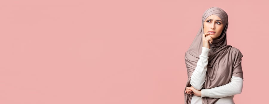 Pensive arabic woman in hijab thinking about something and looking away on pink studio background, panorama with free space photo