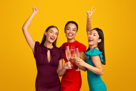 Glamour Party. Three Girls Models In Evening Dresses Clinking Glasses Having Fun Celebrating Something Standing Over Yellow Background photo