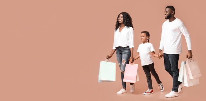 Family Shopping Concept. Happy black parents and their cute little daughter walking with colourful shopping bags over yellow background, panorama photo