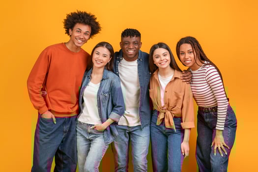 Group of five diverse young adults standing in front of a yellow background. They are all smiling and looking at the camera. The group appears to be friends and is enjoying each others company. photo