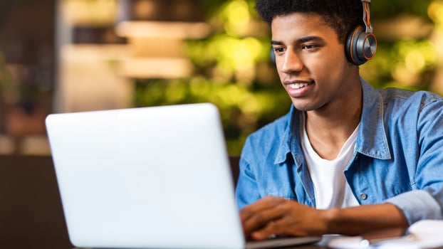 Closeup of African American young guy wearing headphones is focused as he works on a laptop. His hands are typing on the keyboard, and the laptop screen displays graphs and data. photo