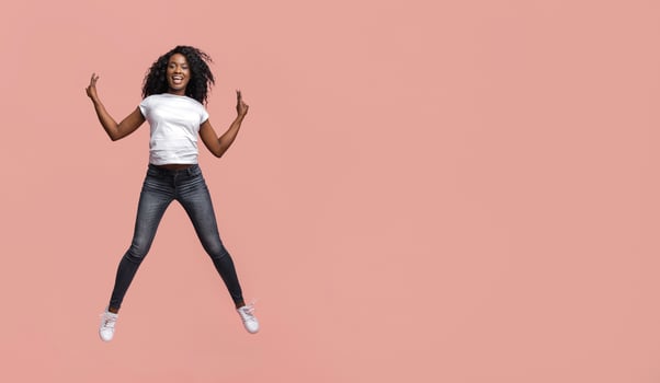 This image shows a young African American woman with curly hair jumping in front of a plain pink background. She is wearing a white t-shirt, dark blue jeans, and white sneakers. photo