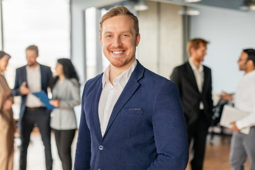 A man in a blue suit and white shirt stands in an office setting, smiling confidently. He is surrounded by colleagues, who are talking and working in the background. The office is bright and modern photo