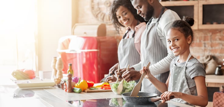 Smiling african little girl helping parents cooking healthy dinner in kitchen, frying vegetables photo