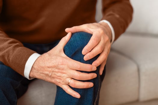 Arthritis concept. Unrecognizable elderly man massaging his knee suffering from pain sitting on couch at home. Selective focus, cropped photo