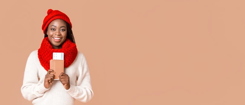 Emotional african woman wearing winter knitted set holding ticket and passport, beige studio background, holiday travelling vacation concept photo