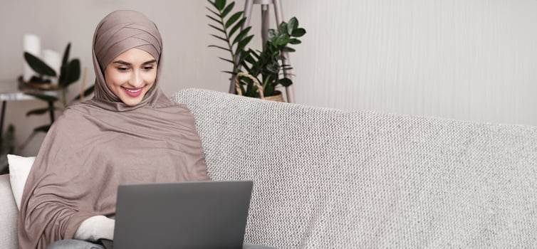Dating Websites For Muslim. Beautiful Arab Girl In Hijab Using Laptop Computer At Home, Chatting Online While Sitting On Cozy Couch, Panorama photo