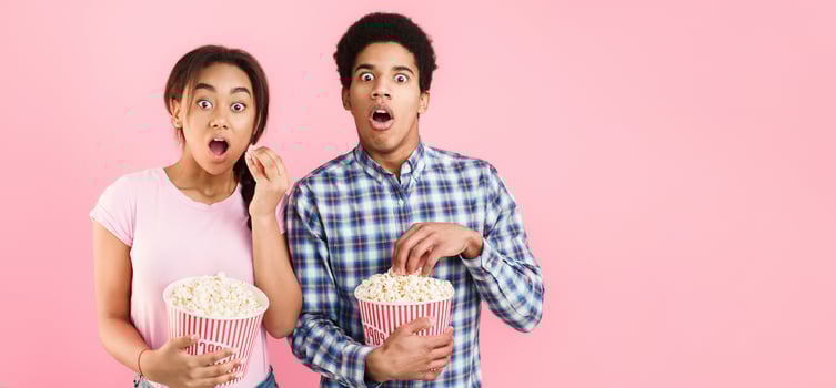 Shocked couple watching scary movie, holding bucket of popcorn on pink background photo