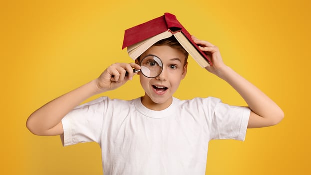 Ready for school. Cute clever boy with magnifying glass in hand hiding under book, orange studio background photo