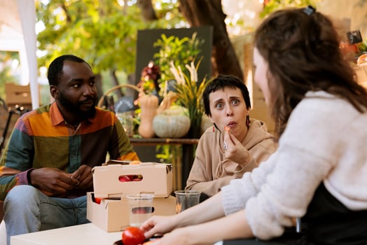 Caucasian farmer offering freshly harvested bio produce to multiracial customers. Young diverse consumers seated and tasting locally grown organic fruits at harvest farm fair festival. photo