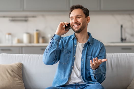 A man with a beard is sitting on a white couch in his home while talking on his phone. He is wearing a blue denim shirt and jeans. He is smiling and gesturing with his hand as he speaks. photo