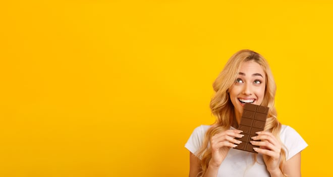 This image shows a young woman smiling and biting into a chocolate bar while looking to her right. She is standing in front of a bright yellow background, blank space photo