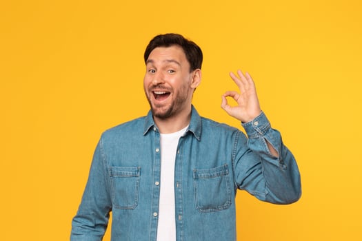 A man with brown hair and a beard, wearing a blue denim shirt over a white t-shirt, smiles while showing the okay gesture with his right hand, standing against a yellow background. photo