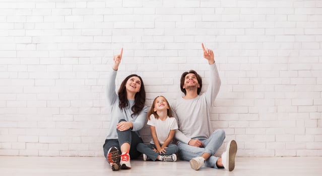 Family of three sitting on floor near white brick wall, pointing and looking upwards, copy space photo