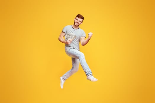 Celebrating Win. Lucky Young Man Jumping On Orange Studio Background With Money In His Hand, Gesturing photo
