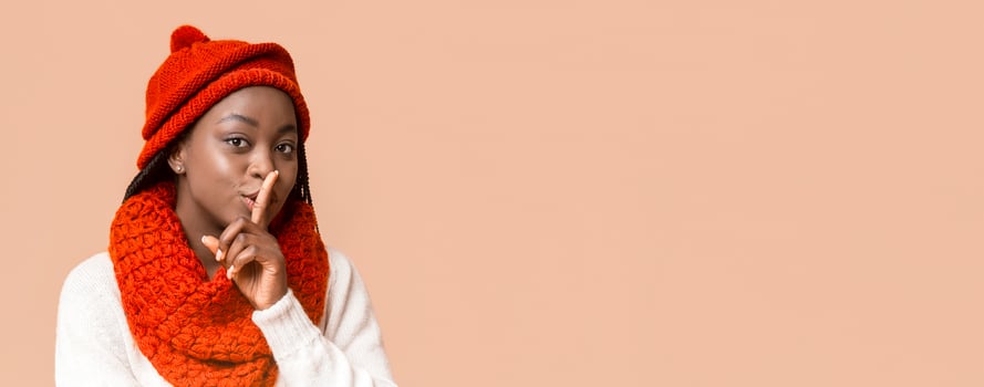 A young black woman softly gestures for silence, wearing a cozy red hat and matching scarf. She holds her finger to her lips, conveying a sense of calm and secrecy, copy space photo