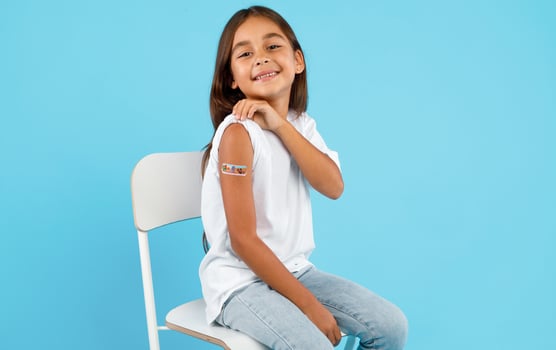 Vaccination Of Children. Happy Vaccinated Kid Girl Showing Arm With Adhesive Bandage After Vaccine Injection Sitting On Blue Studio Background. Kids And Covid-19 Prevention, Antiviral Immunization photo