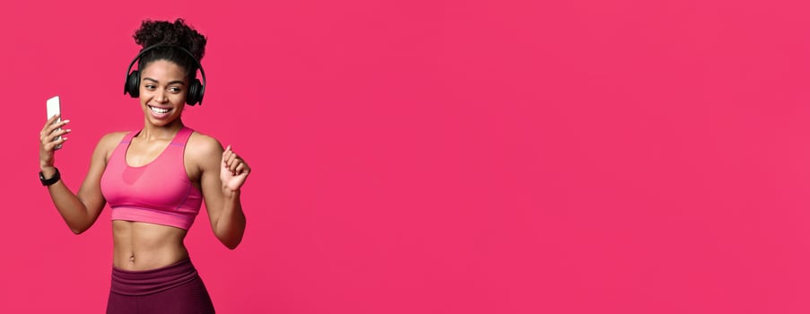 Joyful african fitness girl enjoying music during exercising, holding phone and wearing wireless earphones over red background, sport music application concept, copy space photo