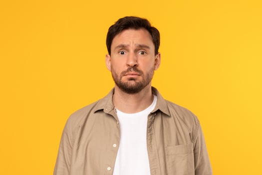 A man with a beard and brown hair is wearing a beige button-up shirt over a white t-shirt. He is looking directly at the camera with a serious expression on his face. The background is a solid yellow. photo