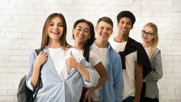 A diverse group of multiethnic teenagers is lined up, displaying a variety of cheerful expressions. The backdrop features a white brick wall, suggesting an informal or educational setting photo