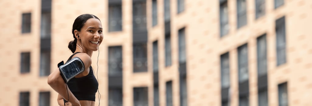 Fit Young Woman In Stylish Sportswear Resting Outdoors After Training, Sporty Athletic Lady With Earphones And Armband Phone Holder Standing On Urban City Street And Smiling At Camera, Copy Space photo