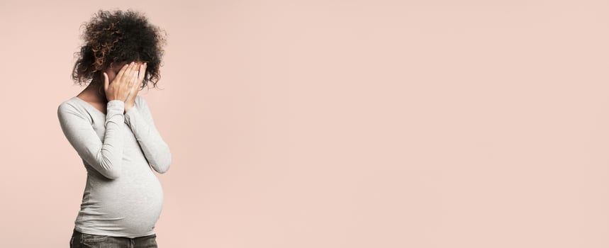 Depression during pregnancy. Afro woman covering face with hands and crying, grey background photo