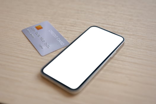A close-up of a credit card and a smartphone with a white screen mockup placed on a wooden table indoors. photo