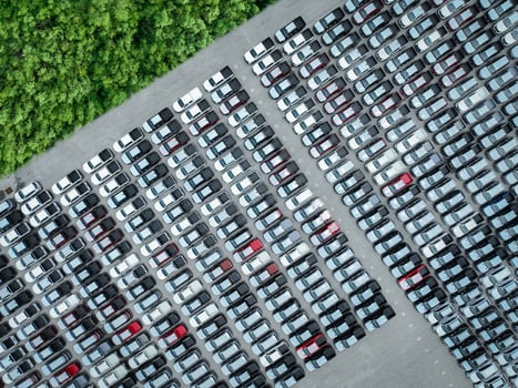 Aerial view of a factory parking lot filled with new cars adjacent to lush green forest. Automotive industry's focus on sustainability, logistics, and zero-emission electric and hybrid vehicles. photo