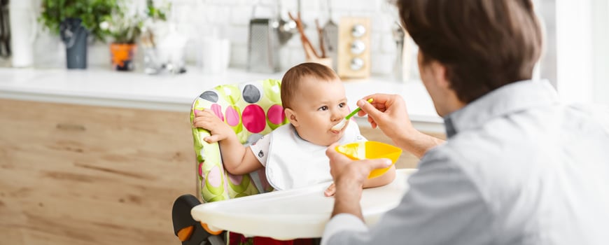 A father feeds his baby in a high chair in a kitchen. The baby is wearing a white bib and is sitting in a colorful high chair. The father is using a spoon to feed the baby, copy space photo