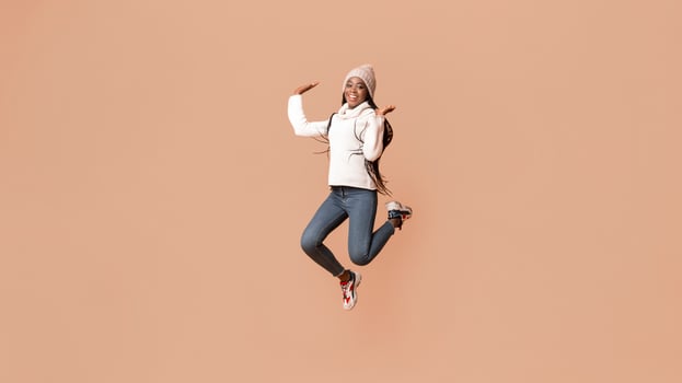 Playful young african woman in winter hat having fun over beige studio background, jumping in the air gracefully photo