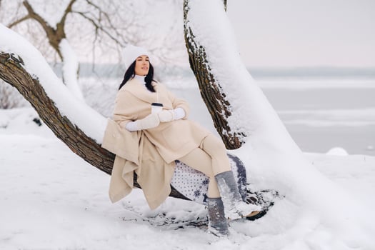 A beautiful girl with a beige cardigan and a white hat enjoying drinking tea in a snowy winter forest near a lake photo
