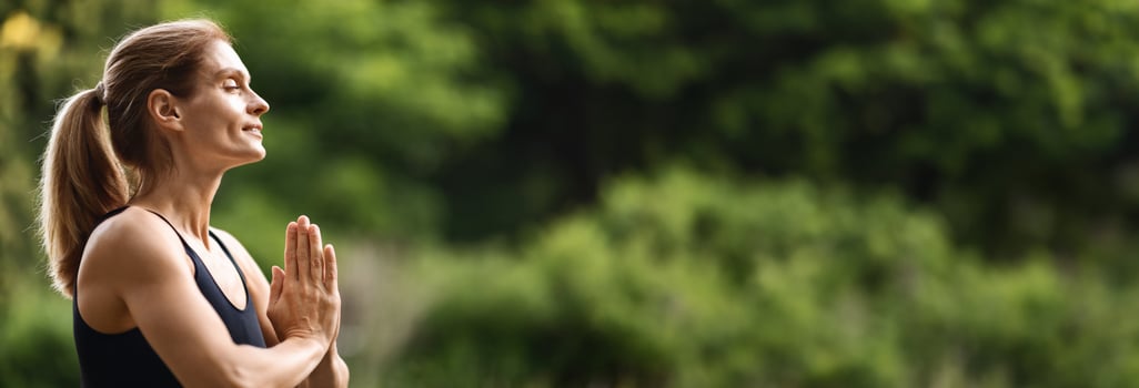 Closeup of happy millennial blonde woman in black tight sportswear meditating alone at public park, holding hands in namaste gesture, eyes closed, side view, panorama with copy space photo
