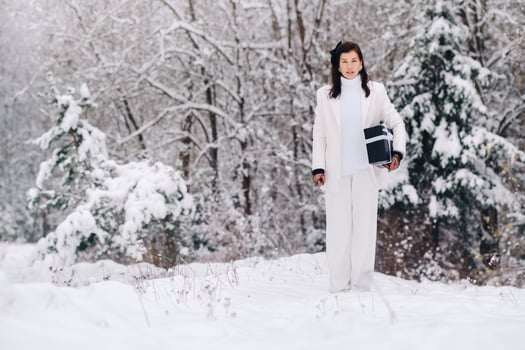 A stylish woman with a white suit with a New Year's gift in her hands in a winter forest. A girl in nature in a snowy forest with a gift box photo