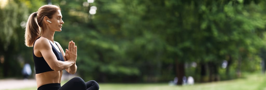 Peaceful millennial blonde woman in black tight sportswear sitting on fitness mat in lotus position with closed eyes, using wireless earbuds, meditating alone at public park, side view, copy space photo