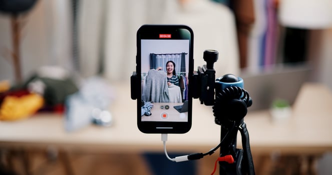 Selective focus on mobile phone of Cheerful woman influencer is live streaming sell clothes on social media or blogging in home studio. Remote buying, Online shopping. photo